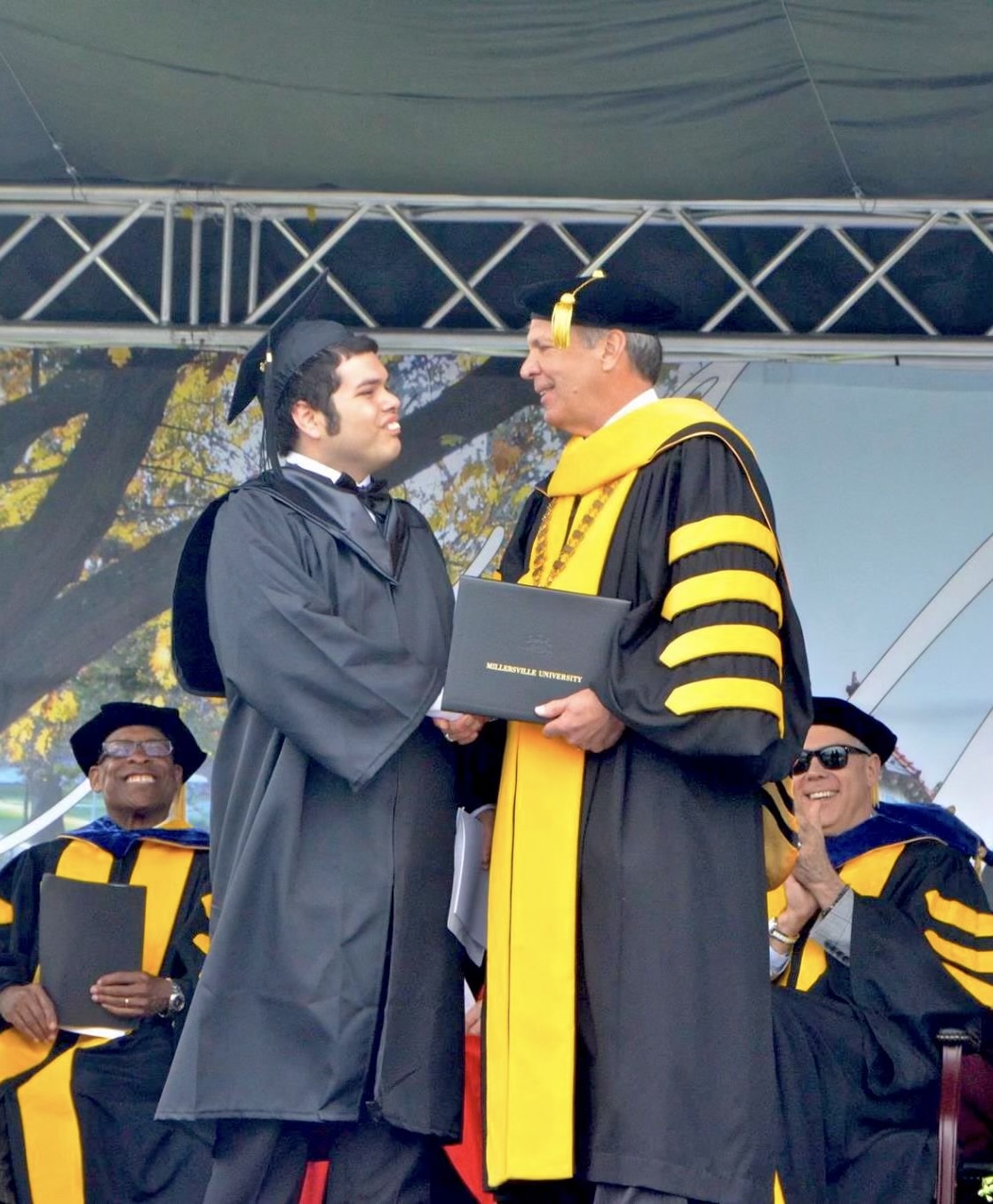 A student wearing black graduation regalia accepts his diploma while shaking hands with a university faculty member.